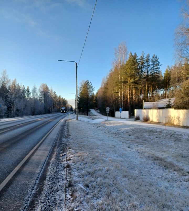 Jäätynyt maantie ja vaalea nurmikko aurinkoisena talvipäivänä; taustalla metsää ja taloja.