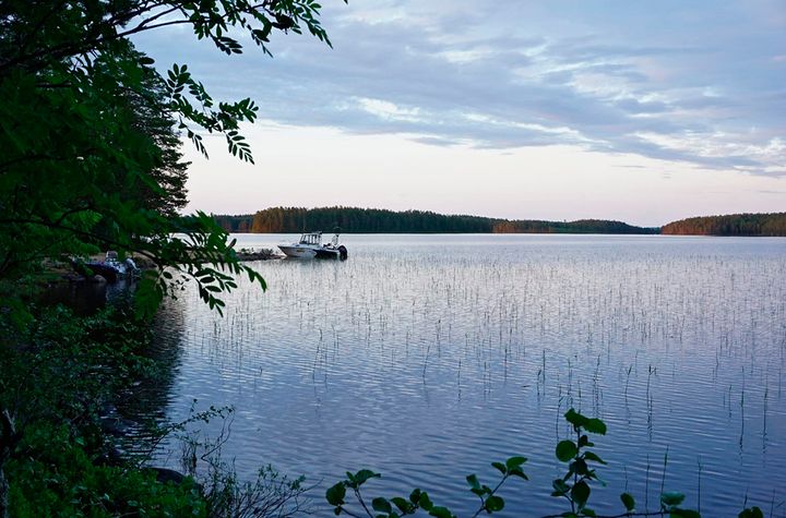 Kesäinen ilta vesistön äärellä, edustalla ranta ja laituri, johon kiinnitettynä moottorivene.