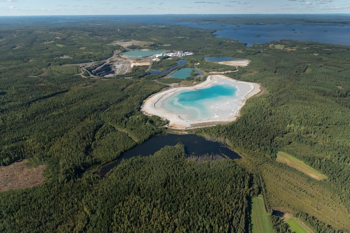 Ilmakuva Elementis Mineralsin Sotkamon tehdas- ja kaivosalueesta, etualalla näkyy laajoja vihreitä metsäalueita ja vaaleampi rikastushiekka-allas, taustalla järvi ja sininen taivas.