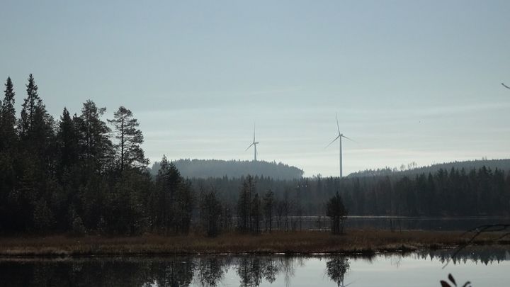 Kaksi tuulivoimalaa metsä- ja järvimaisemassa.