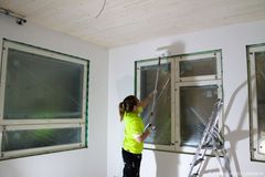 Henkilö maalaa kattoa valkoiseksi huoneessa, jossa on suuret muovitut ikkunat.