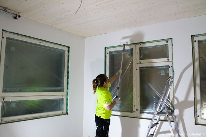 Henkilö maalaa kattoa valkoiseksi huoneessa, jossa on suuret muovitut ikkunat.