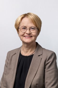 Kuvassa on hymyilevä henkilö pukeutuneena tummaan puseroon ja vaaleaan jakkutakkiin.