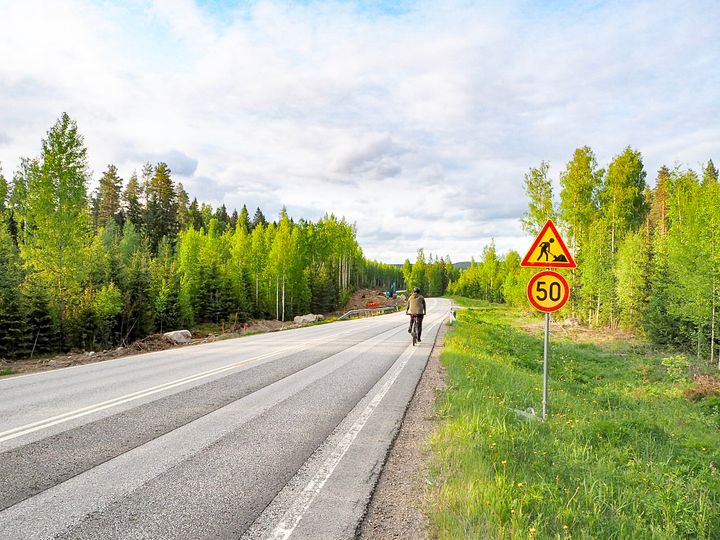 Pyöräilijä polkemassa kamerasta pois päin maantiellä. Tien vieressä kyltti tietyömaasta sekä 50 km/h nopeusrajoitus. Tien toisella puolella työmaa, jossa tehdään kevyen liikenteen väylää.