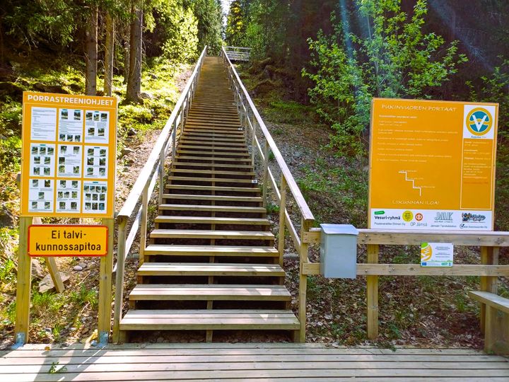 Puiset kuntoportaat, jotka nousevat ylös metsäistä rinnettä. Portaiden alapäässä kaski infokylttiä.