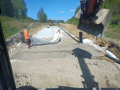 Kaksi työntekijää levittää valkoista geotekstiiliä soratien päälle työmaalla. Taustalla näkyy metsämaisema ja kirkas sininen taivas.