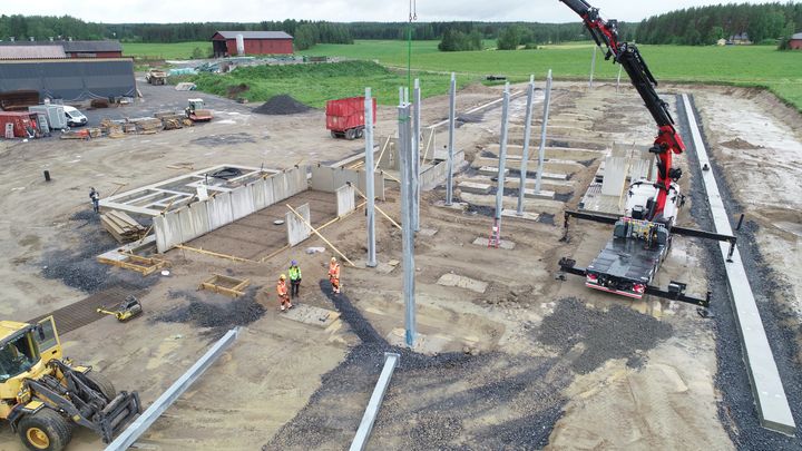 Iso rakennustyömaa alkamassa, betonikehikot ja betonipilarit valettu, muutama ihminen suojakypärät päässä ja taustalla peltomaisemaa.