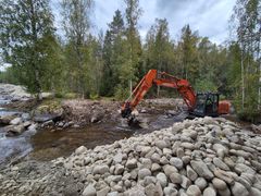 Kaivinkone työskentelee joen varrella kivisessä maastossa.