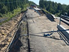 Vaikeasti vahingoittunut tieosuus metsäalueella, jossa kaide on vääntynyt ja osa asfaltista romahtanut. Taustalla työskentelee työntekijöitä.
