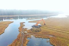 Kaivinkone työskentelee suolla, joka ympäröi vesistöä. Taustalla metsää ja sumua.