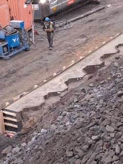 Rakennustyömaa, jossa työntekijä kävelee kypärä päässä. Etualalla sorakasa ja keskeneräinen betonimuotti.