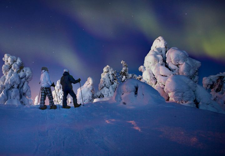 Kuvassa kaksi lumikenkäilijää lumisessa maisemassa, taivaalla revontulia.