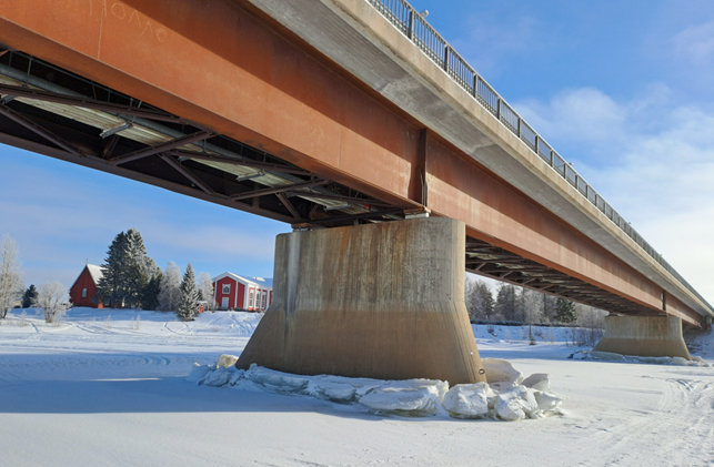 Alhaalta kuvattu silta, joki jäässä, taustalla taloja.
