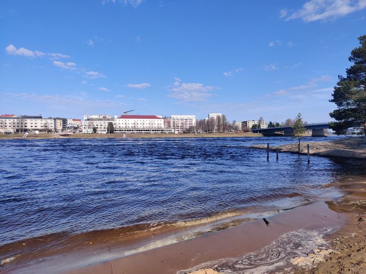 Joki virtaa hiekkarannan vieressä. Taustalla on kaupunki.
