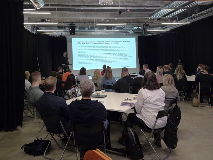 Ihmisiä selin kameraan katselemassa lavalla näytettävää PowerPoint-esitystä.
