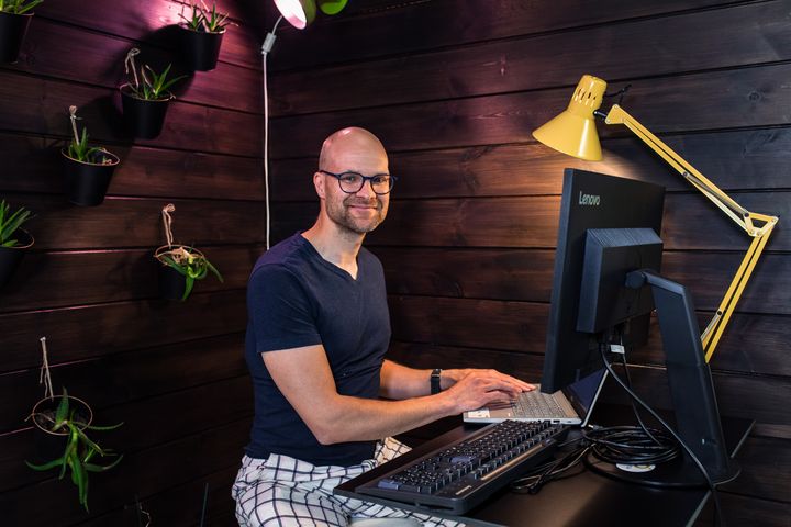 Mies istuu tietokoneen ääressä ja katsoo hymyillen kameraan.