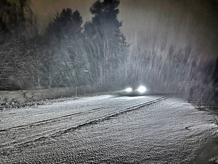 Talvinen lumisade pimeällä, auton etuvalot loistavat pimeässä.