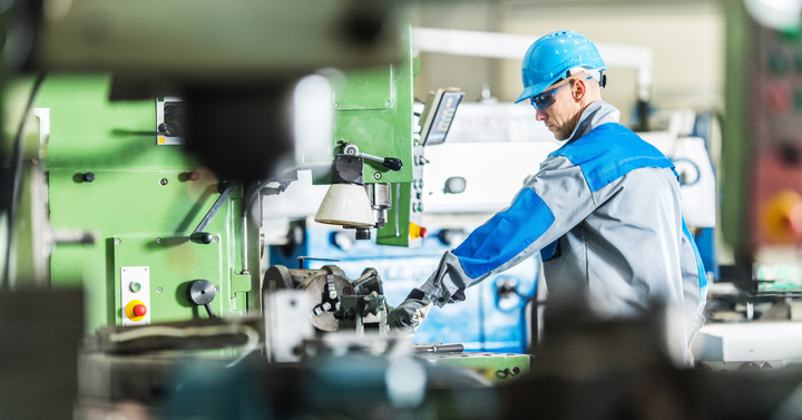 Tämä on valokuva tehdastyöntekijästä, joka käyttää vihreää konetta. Työntekijä on pukeutunut siniseen kypärään ja siniseen työasuun. Kone on pora- tai jyrsinkone, jossa on nappeja ja vipuja. Taustalla näkyy muita koneita ja laitteita tehtaalla.