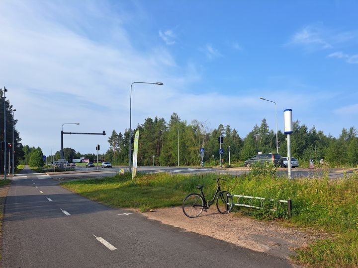 Valtatien risteysalue, jossa jalankulku- ja pyörätie, polkupyörä telineessä, liikennevalot, automaattinen valvontakamera ja autoja.