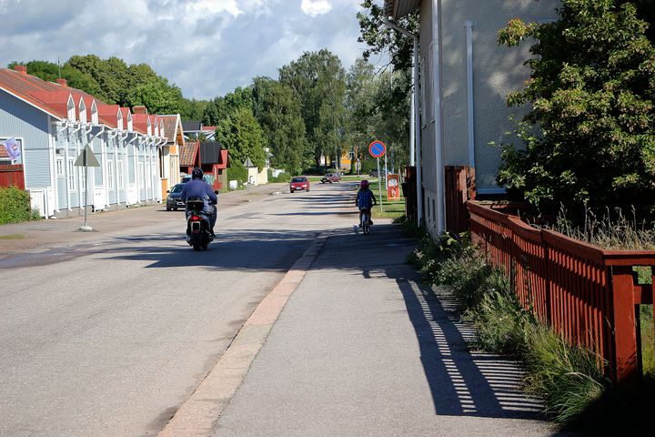 Pieni katu. Lapsi ajaa polkupyörällä ja mies ajaa mopedilla.