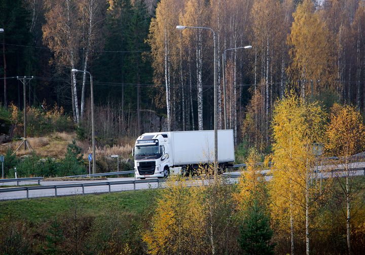 Valkoinen kuorma-auto ajaa syksyisessä maisemassa, taustalla näkyy keltaisia ja ruskeita puiden lehtiä.