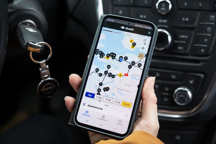 Käsi, joka pitää puhelinta autossa. Puhelimen näytöllä on auki Fintraffic Mobiili -sovellus.