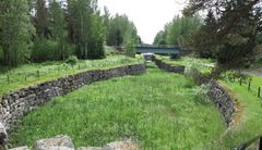 Kuva Taipaleen vanhasta kanavasta Varkaudessa, kivimuurein reunustettu ja kasvillisuuden peittämä kanava. Taustalla silta ja metsää.