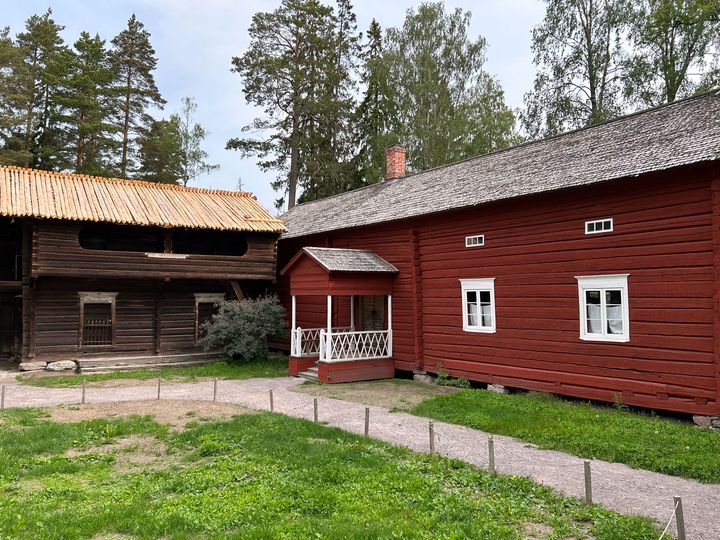 Punainen perinteinen puutalo ja vanha puinen rakennus kesäisessä pihapiirissä.