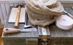 Puulaatikon päällä on käsityövälineitä, kuten vasara, talttoja, ja rulla pakkausmateriaalia.