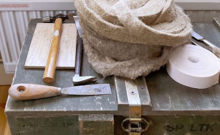 Puulaatikon päällä on käsityövälineitä, kuten vasara, talttoja, ja rulla pakkausmateriaalia.