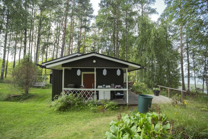 Pieni tummanruskea mökki järven rannalla vehreässä maisemassa.