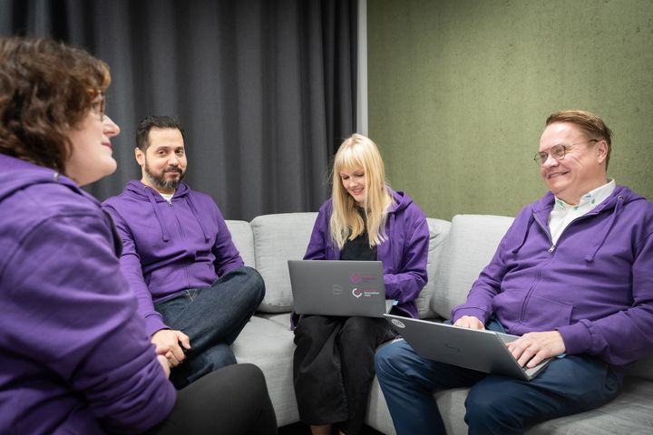 Neljä henkilöä istuu sohvalla pukeutuneena violetteihin huppareihin ja työskentelee kannettavilla tietokoneilla.