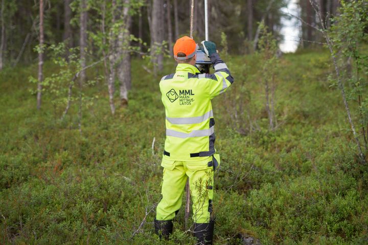 Lantmäteriingenjören står med ryggen mot kameran i gula skyddskläder och tittar på skärmen på sin enhet för att fastställa gränsen i skogen.