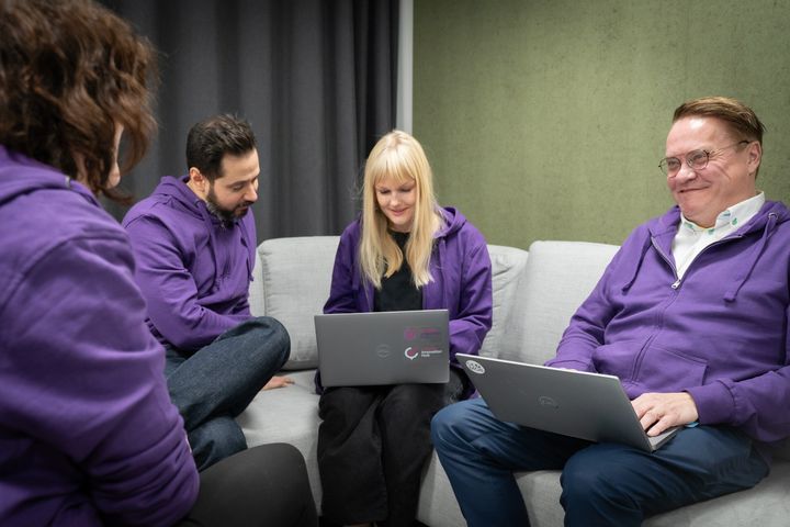 Fyra personer sitter, två har datorer, alla har lila luvtröjor på sig.