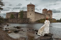 Valkopukuinen nainen seisoo rannalla ja katsoo Olavinlinnaa.
