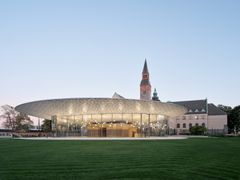 Kansallismuseon valaistu paviljonki ja tornillinen päärakennus iltahämärässä, edessä nurmikenttä.