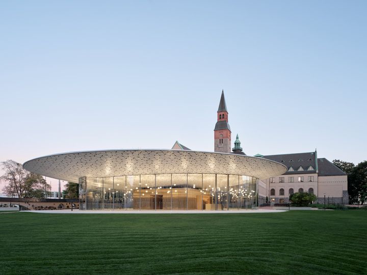 Kansallismuseon valaistu paviljonki ja tornillinen päärakennus iltahämärässä, edessä nurmikenttä.