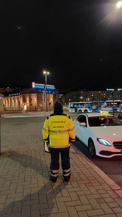 Työsuojelutarkastaja seisoo taksin vieressä, taustalla Hakaniemen kauppahalli.