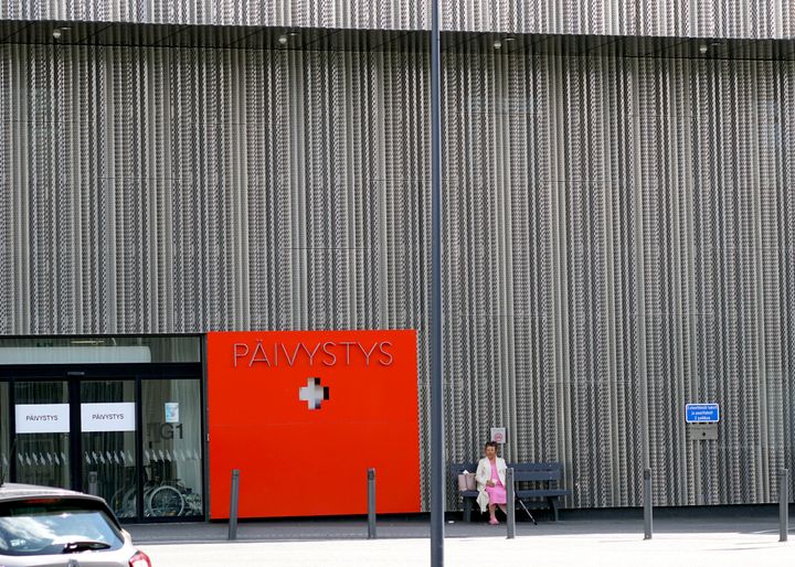 Punainen päivystyksen kyltti rakennuksen ulkopuolella, nainen istuu penkillä.