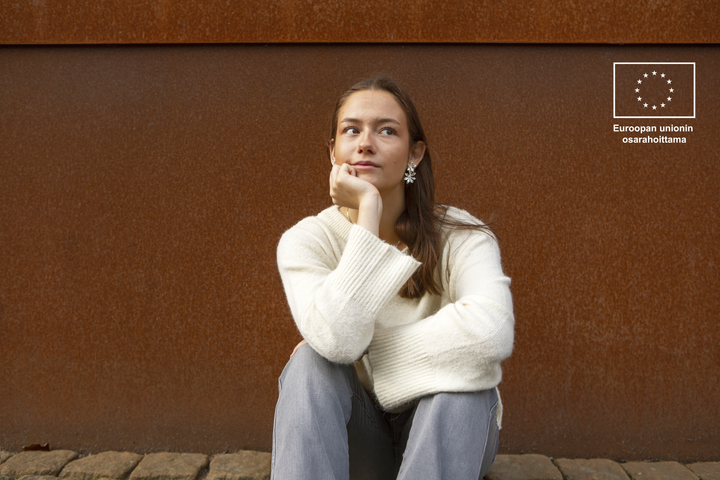 Henkilö istuu ulkona ja katsoo ylös mietteliäänä. Taustalla ruosteenvärinen seinä ja EU-logo.