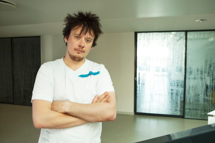 Hoitajan asussa oleva miesoletettu henkilö seisoo sisätilassa Diakin Helsingin-kampuksella, kädet ristittyinä. Taustalla on lasiovia.