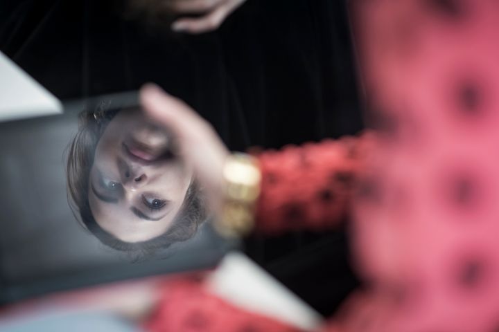 Etualalla näkyy sumuinen henkilön olkapää. Kameran tarkennus on sammutetussa näytössä, joka heijastaa nuoren naisen kasvot.