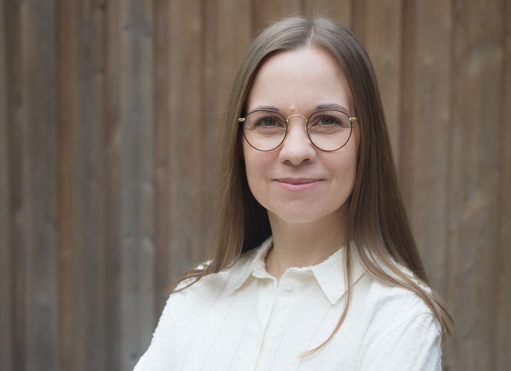 Aura Vuorenrinne seisoo puisen seinän edessä ja katsoo kameraan. Hänellä on pitkät hiukset ja silmälasit, ja hän on pukeutunut vaaleaan puseroon.