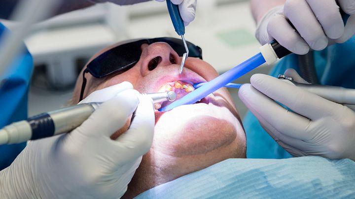 Hammaslääkäri hoitaa potilasta, jolla on aurinkolasit päässä, ja käyttää erilaisia hammashoitovälineitä.