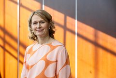 Nainen seisoo värikkäässä taustassa päällään oranssi ja vaaleanpunainen kuvioitu paita.