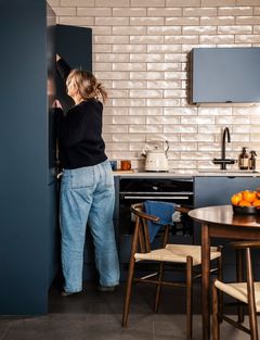 Sinisen keittiö, jossa naisoletettu kurottaa aamiaiskaapin ylähyllylle.