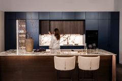 Keittö, jossa tyrskyn siniset ja Agostino tammen sävyiset kaapinovet, saareke ja kaksi vaaleaa baarijakkaraan. Nainen on avaamassa kaapinovea.