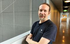 Siniseen Helsingin yliopiston tietojenkäsittelytieteen osaston T-paitaan pukeutunut parrakas, hymyilevä mies nojaa kaiteeseen. Taustalla on käytävä ja metalliristikkoseinä.