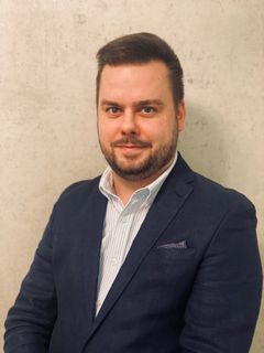 Henkilö istuu vaalean taustan edessä pukeutuneena tummaan pukuun ja raidalliseen kauluspaitaan.