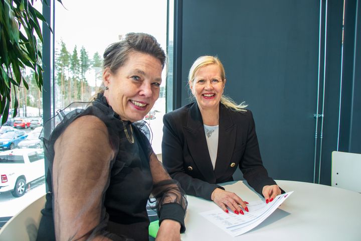 Kaksi naista istuu pöydän ääressä keskustelemassa. Taustalla ikkuna, josta näkyy parkkipaikka.